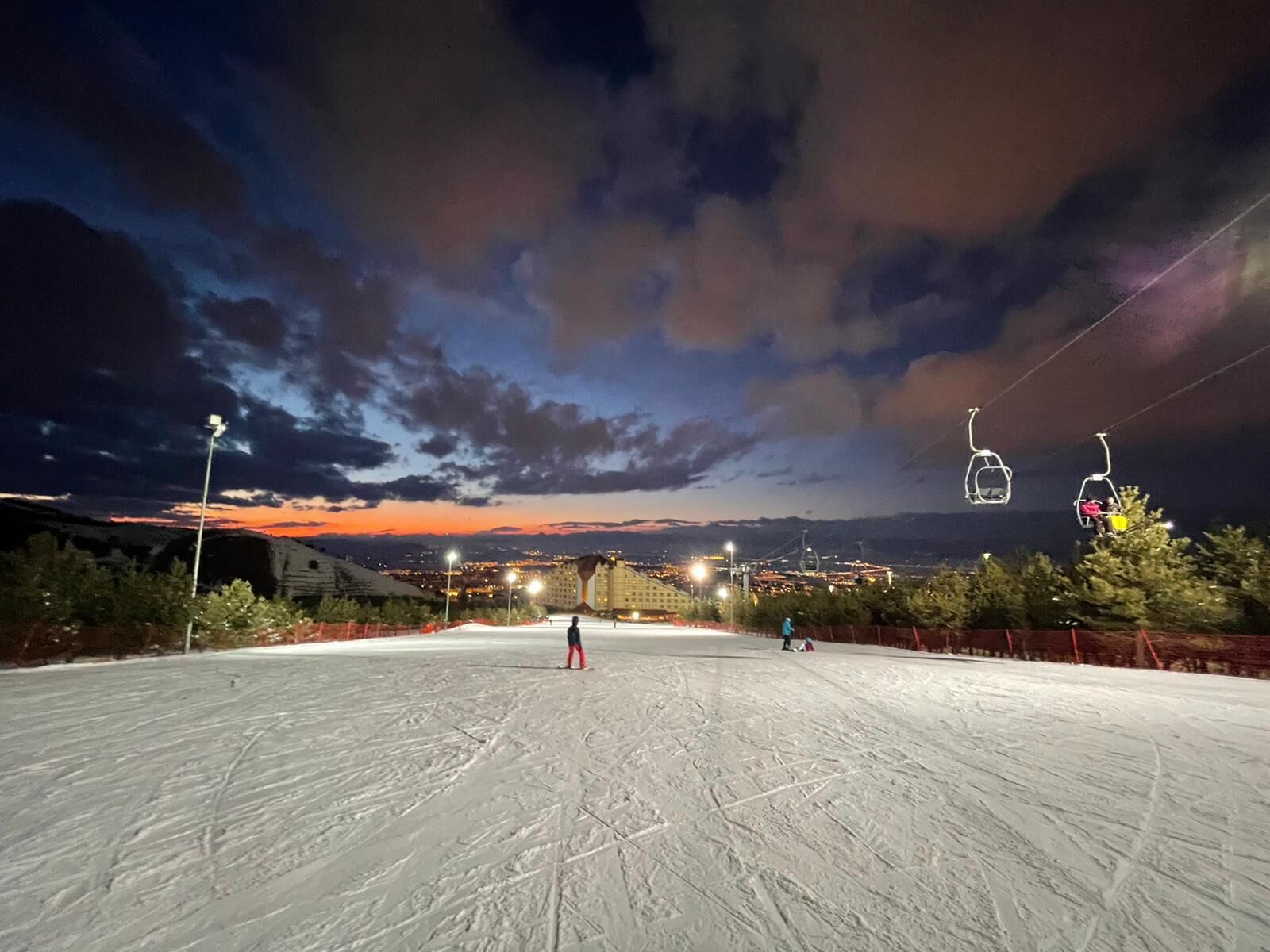 A ski slope at dusk with ski lifts and a distant cityscape.