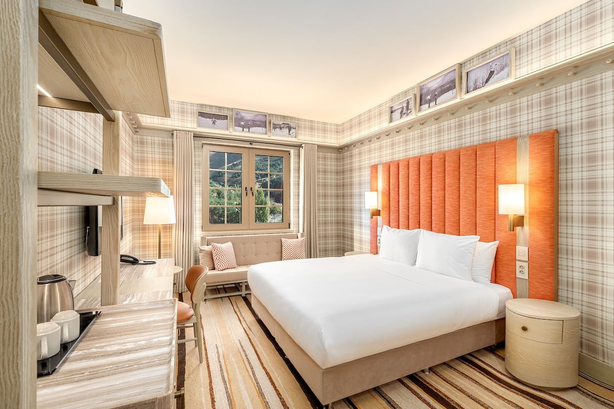 A hotel room with a large bed, a bedside table, a lamp, a window with a view of greenery, and a patterned wall.