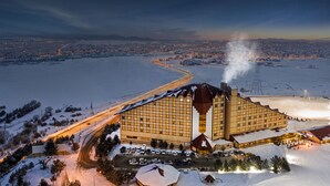 A large hotel with a distinctive architectural design, situated in a snowy landscape with a view of a river and distant city lights. - Polat Palandoken (Erzurum)