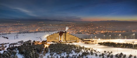 Un gran hotel con una estructura de techo distintiva, rodeado por un paisaje nevado y árboles, con un paisaje urbano al fondo.