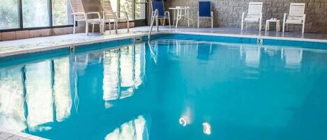 En indendørs swimmingpool med klart, blåt vand, omgivet af hvide liggestole og en murstensmur.