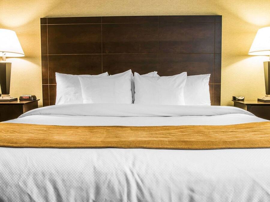 Een netjes opgemaakt bed met witte lakens en een houten hoofdbord, met aan weerszijden nachtkastjes, waarvan er één een lamp en een klok heeft.