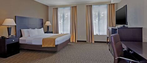 Habitación de hotel con una cama, dos mesitas de noche con lámparas, un televisor sobre un mueble bajo, un escritorio con una silla y ventanas grandes con cortinas.