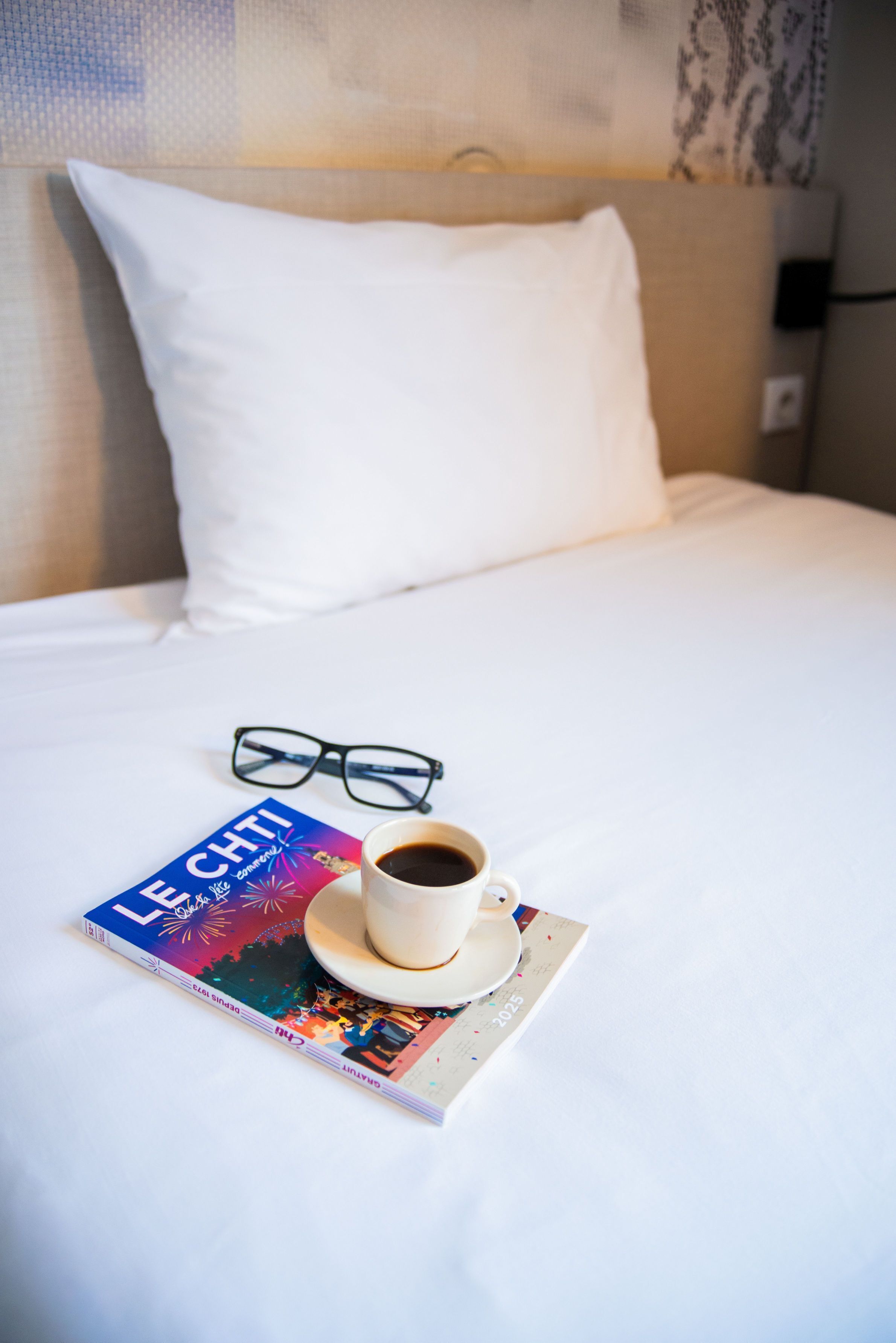A neatly made bed with a pillow, a pair of glasses, a cup of coffee, and a travel magazine on a white bedspread.