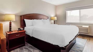 A hotel room with a large bed, two bedside tables with lamps, a window with blinds, and an air conditioning unit.