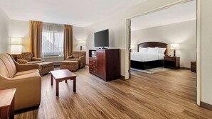 Habitación de hotel con sofá, sillón, mesa de centro de madera, televisión, cómoda y cama con mesita de noche.
