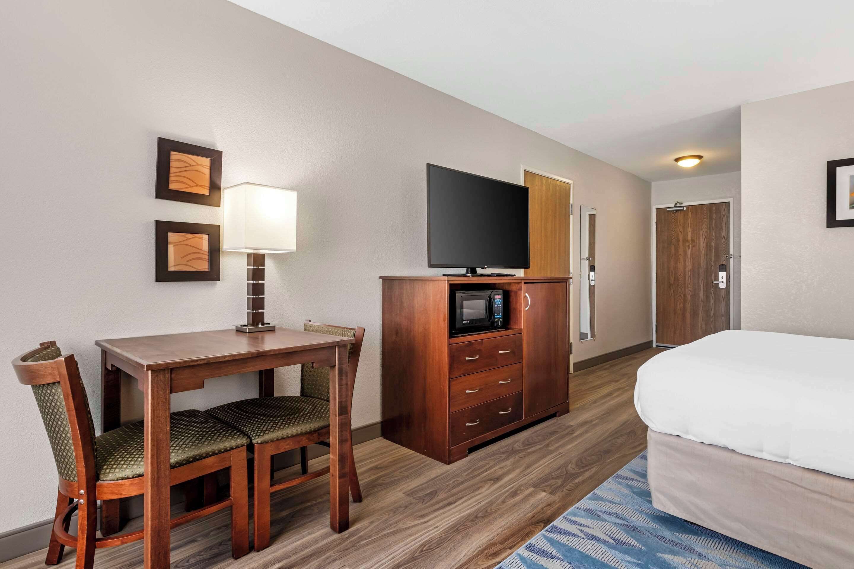 A hotel room with a wooden desk, two chairs, a television, a microwave, a bed, and a door.