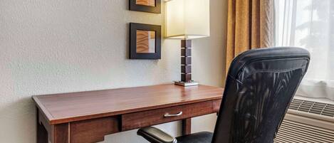 A hotel room with a wooden desk, a black office chair, a wall-mounted lamp, and two framed pictures.