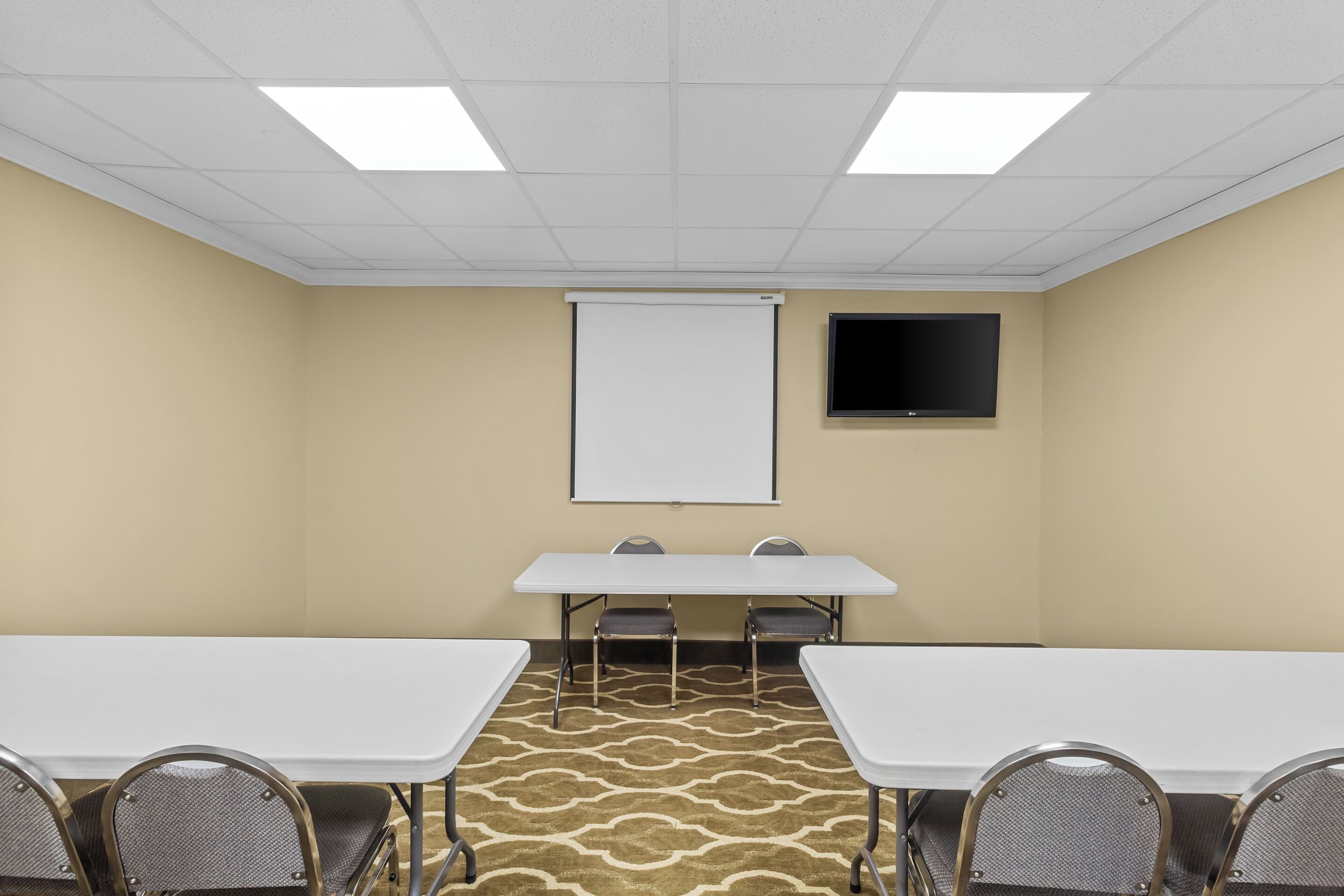 Une salle de conférence avec une table blanche, des chaises, un écran de projection et un téléviseur à écran plat fixé au mur.