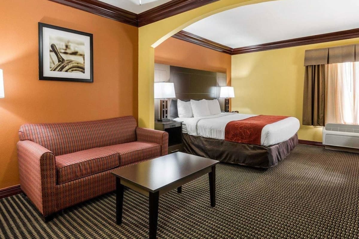 Ein Hotelzimmer mit Bett, Sofa, Tisch, Deckenventilator, gerahmtem Bild, Lampe und einem Fenster mit Vorhängen.