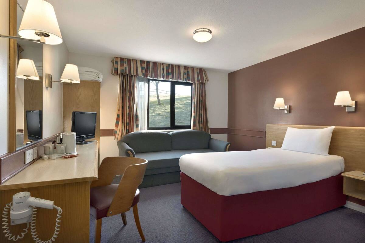 A hotel room with a bed, a desk with a telephone, a chair, a sofa, a television, and a window with curtains.