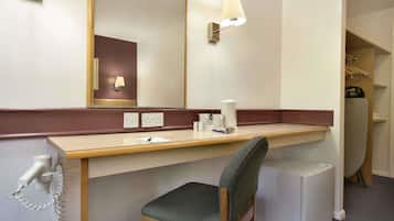 Quarto de hotel com escrivaninha, cadeira, espelho e um pequeno frigorífico.