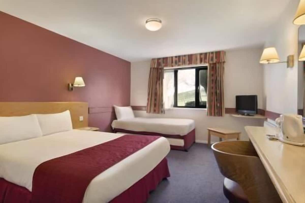 A hotel room with a large bed, a smaller bed, a desk with a chair, a television, and a window with curtains.