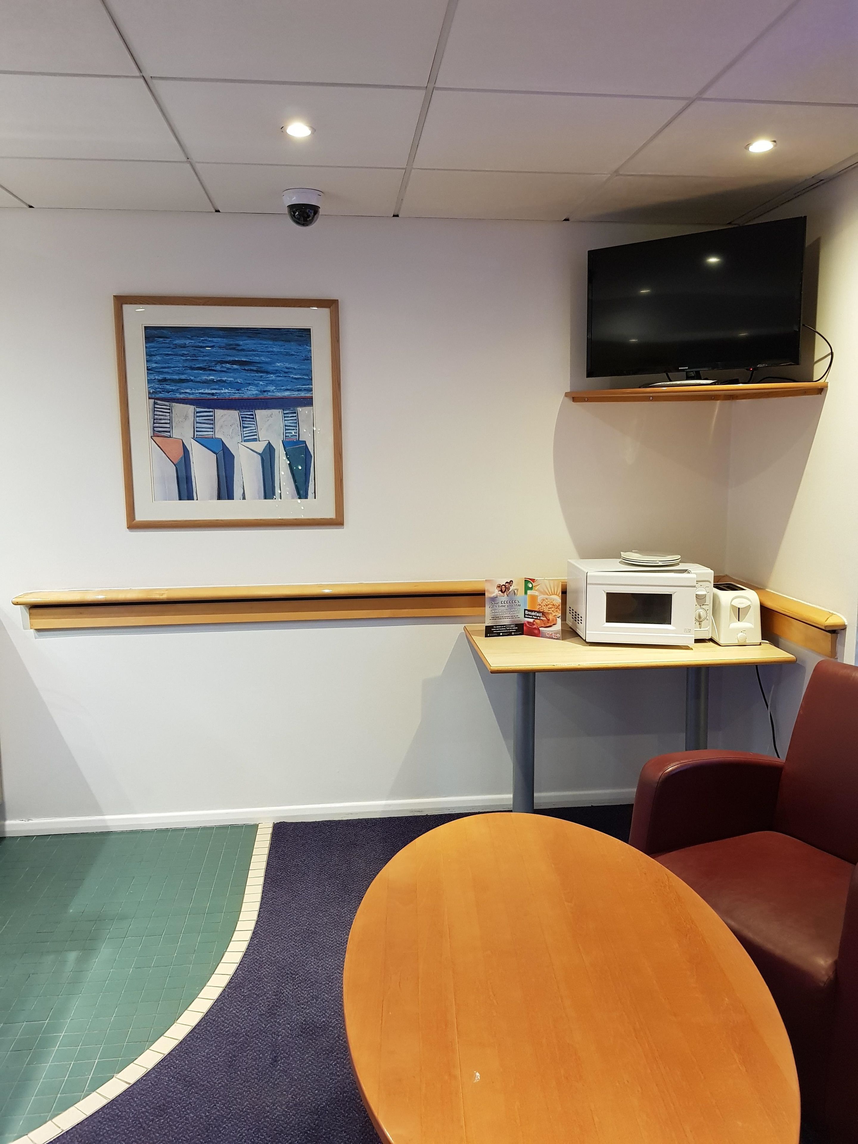 A hotel room with a wooden bench, a table with a microwave and toaster, a TV mounted on the wall, and a framed artwork depicting abstract figures.