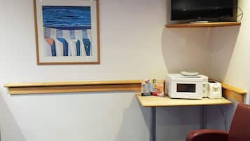 A hotel room with a wooden bench, a table with a microwave and toaster, a TV mounted on the wall, and a framed artwork depicting abstract figures.