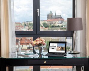 A laptop on a glass desk with a vase of flowers, a cup, and a book. A window reveals a cityscape with a church. - Four Seasons Hotel Prague (Prague)