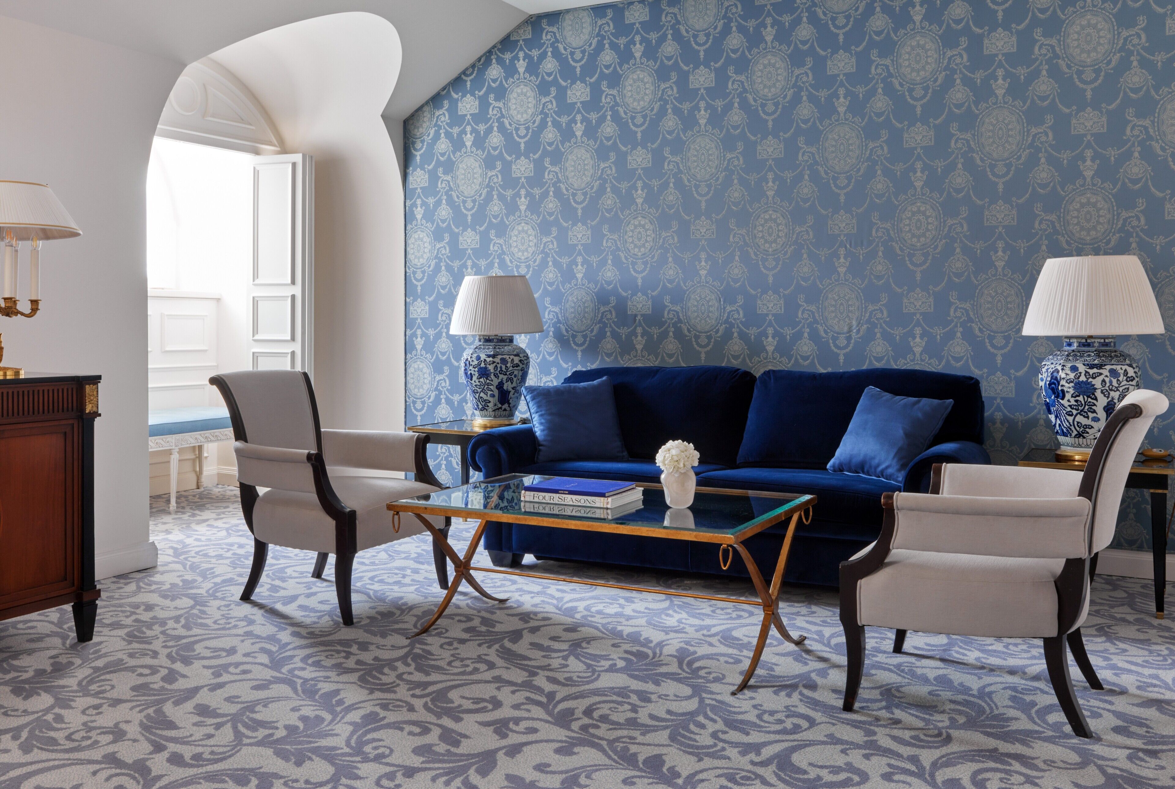 A living room with a blue patterned wallpaper, a blue sofa, two armchairs, a glass coffee table, and two lamps.