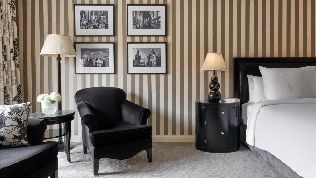Camera d'albergo con una poltrona in pelle nera, un tavolino rotondo con una lampada, un comodino nero con una lampada, un letto con lenzuola bianche e carta da parati a righe con quadri appesi.