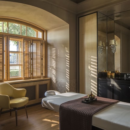A bedroom with a bed, a chair, a large window with shutters, a mirror, and a dresser with a lamp.