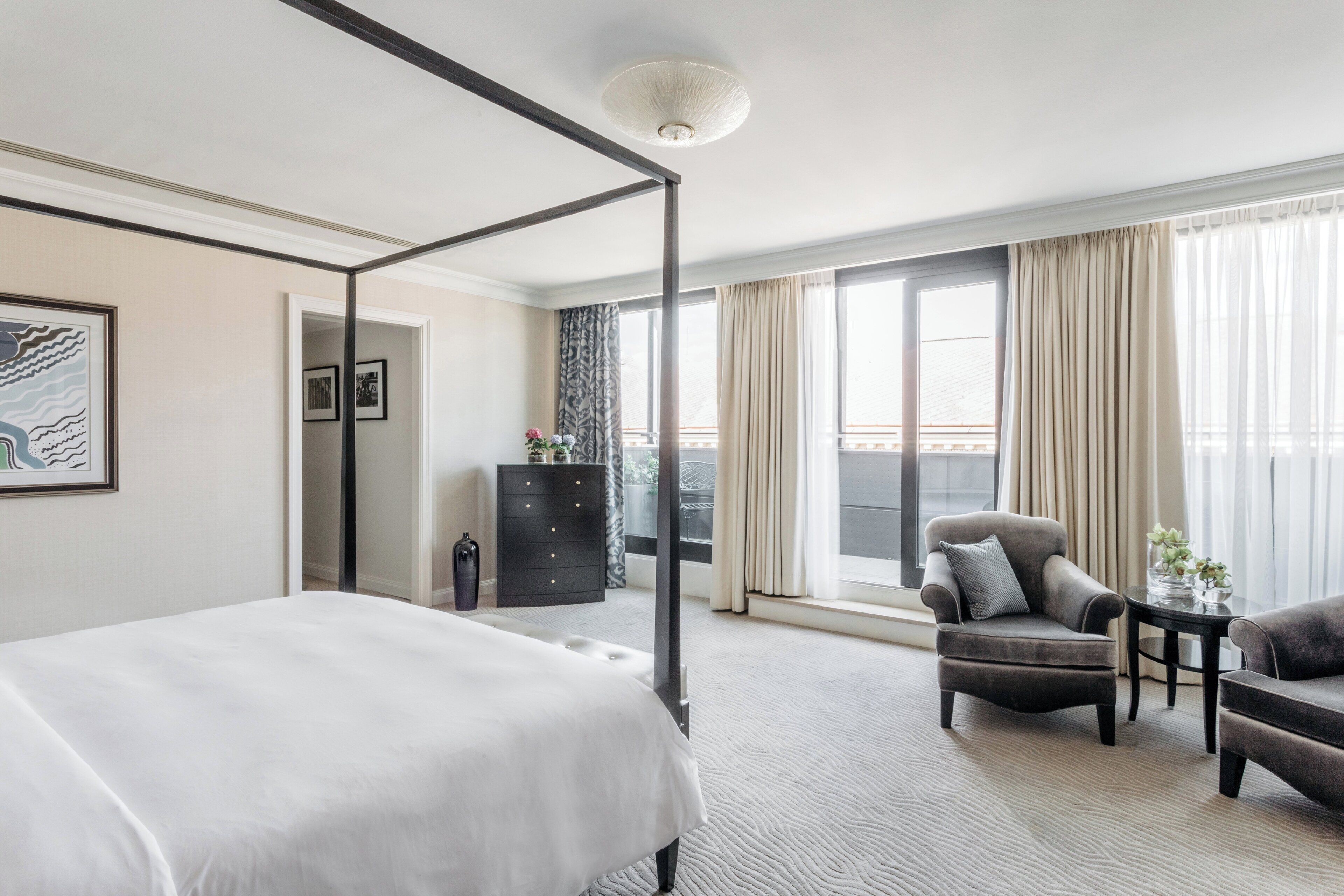 Une chambre d’hôtel moderne avec un lit à baldaquin, un coin salon comprenant un fauteuil et une table, une commode et une grande fenêtre avec des rideaux.