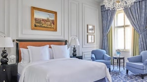 A hotel room with a large bed, two armchairs, a chandelier, and a painting on the wall.