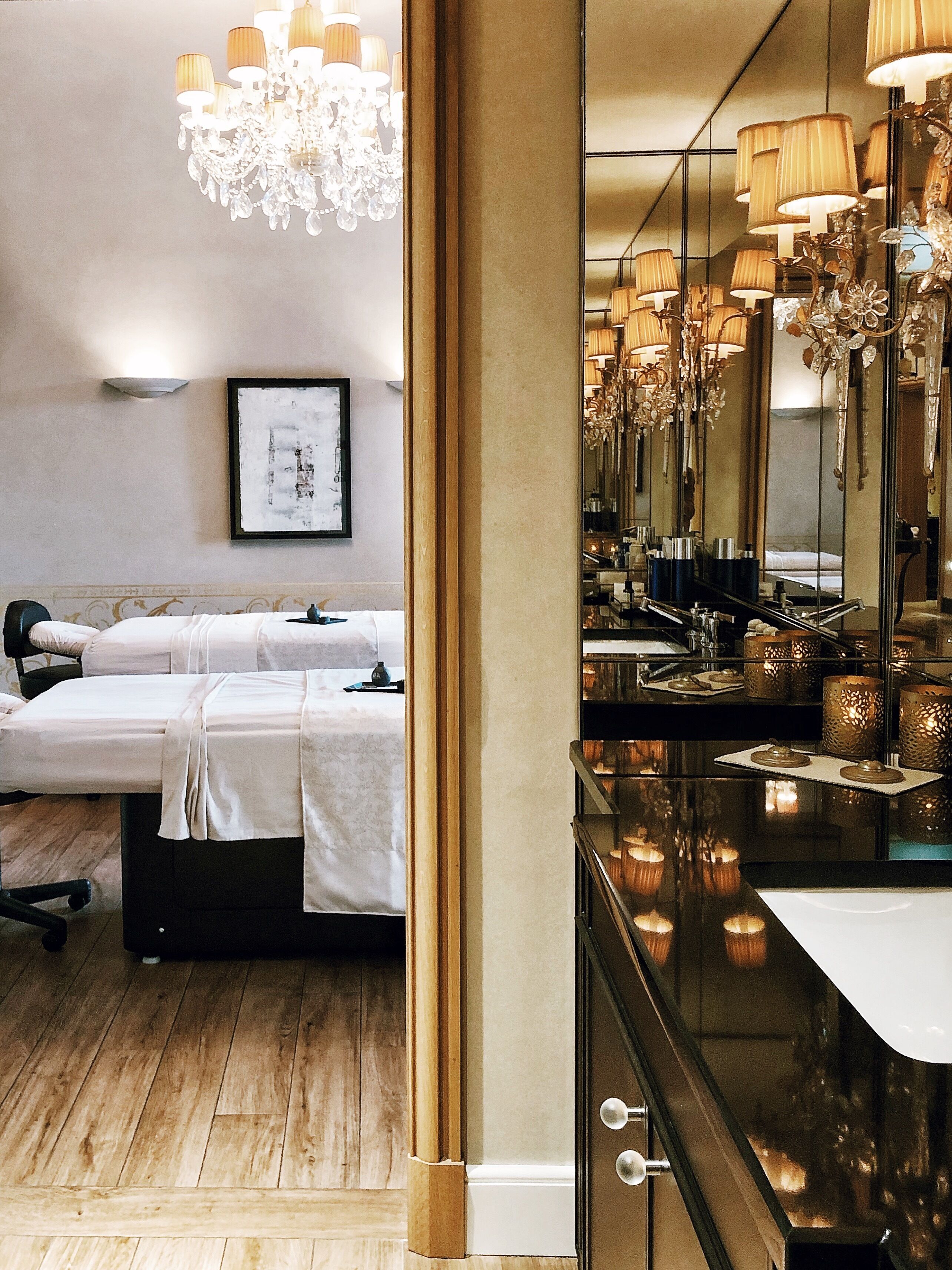 Een hotelkamer met een bed, nachtkastje en een grote spiegel die een kroonluchter weerspiegelt.