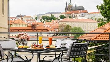 Un balcon avec une table préparée pour le petit-déjeuner, comprenant des fruits et du jus, donnant sur un paysage urbain avec des bâtiments historiques.