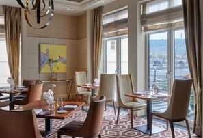 A hotel dining area with multiple tables and chairs, large windows with a view of a bridge and cityscape, and a modern chandelier.