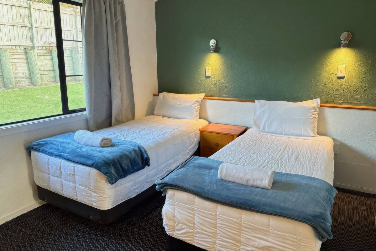 A hotel room with two beds, a window with curtains, and a green wall with wall-mounted lights.