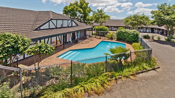 A pool area with a fenced swimming pool, surrounded by a garden with various plants and trees. - Oakwood Manor Motor Lodge (Auckland)