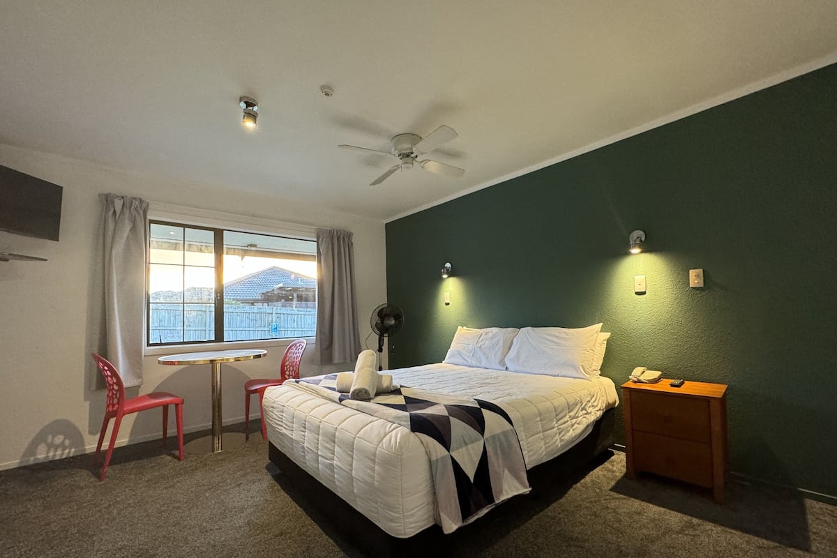 A hotel room with a bed, a desk, a chair, a ceiling fan, and a window with curtains.