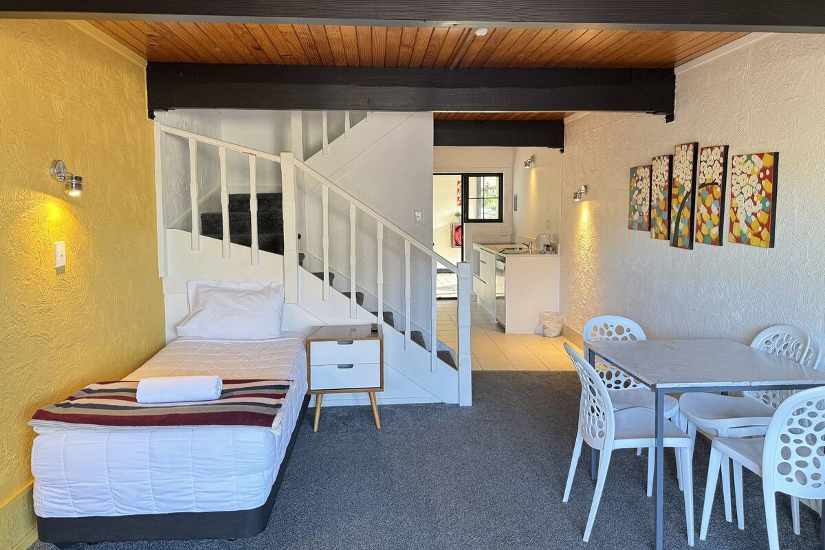 A bedroom with a bed, a small bedside table, a white chair, a dining table with chairs, a staircase, and a kitchen area.