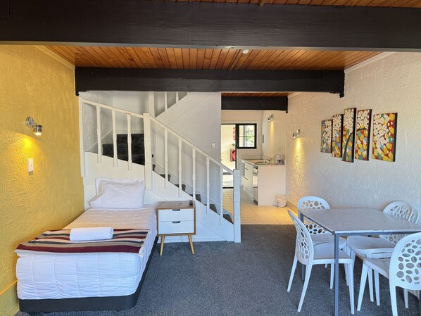 Un dormitorio con cama, una mesita de noche pequeña, una silla blanca, una mesa de comedor con sillas, una escalera y una zona de cocina.