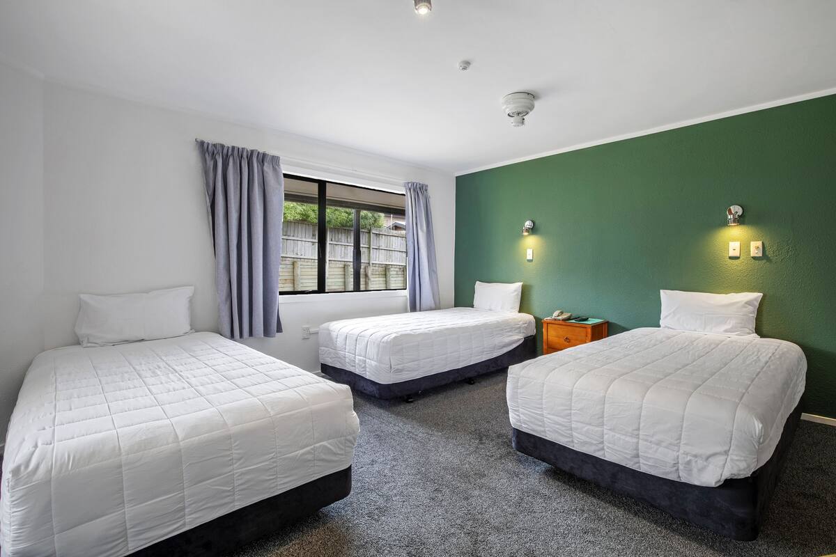 A room with three single beds, a window with curtains, a green wall, and two wall-mounted light switches.
