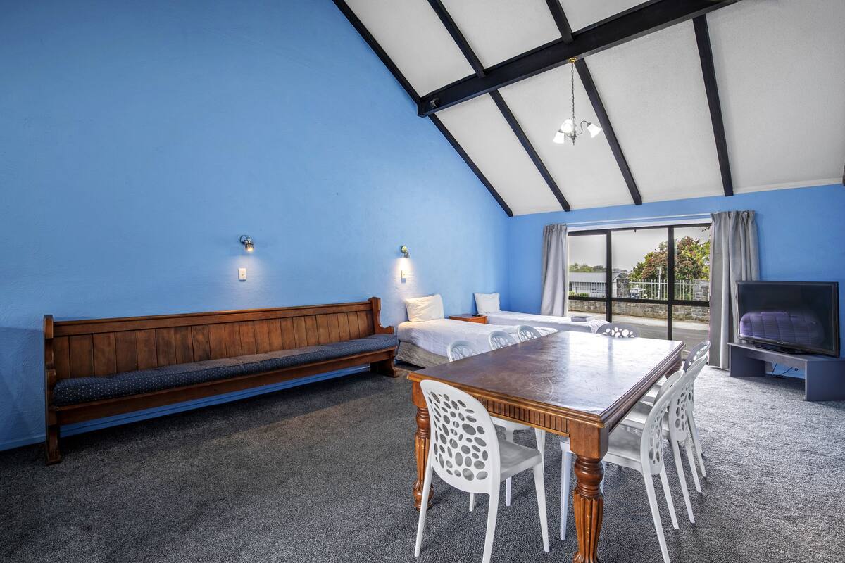 A room with a wooden bench, a dining table with chairs, a bed, and a TV.