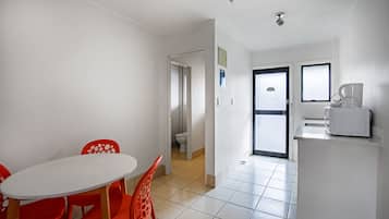 Una habitación pequeña y limpia con una mesa blanca y sillas rojas, una kitchenette y una puerta que da a otra habitación.