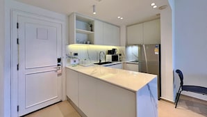 A modern kitchen with white cabinetry, a built-in island, a stainless steel refrigerator, and a black chair. - Allamanda Laguna Phuket by RESAVA (Choeng Thale)