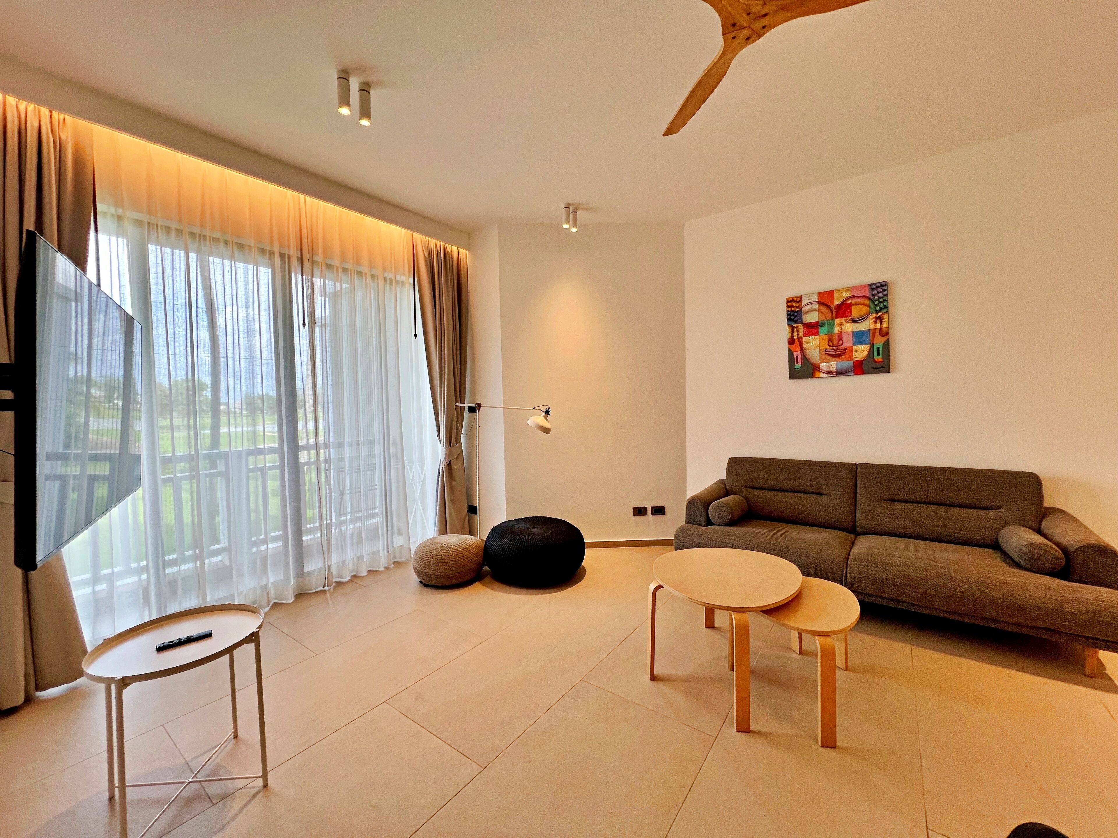 A modern living room with a sofa, two small tables, a bean bag, and a large window with sheer curtains.