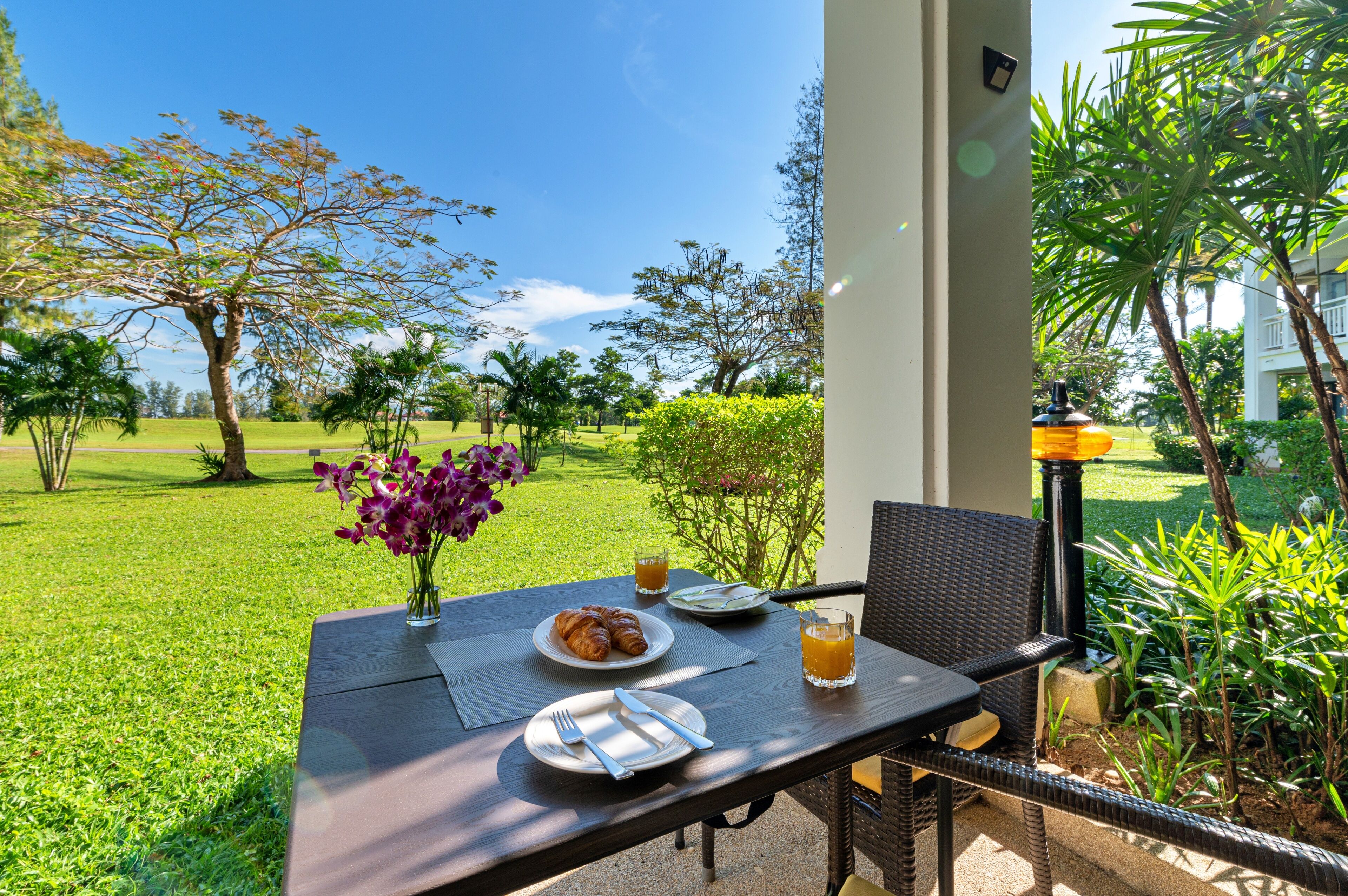 Un espace de restauration en plein air avec une table dressée pour le petit-déjeuner, comprenant des croissants et du jus de fruits, le tout entouré d’une végétation luxuriante et sous un ciel bleu dégagé.