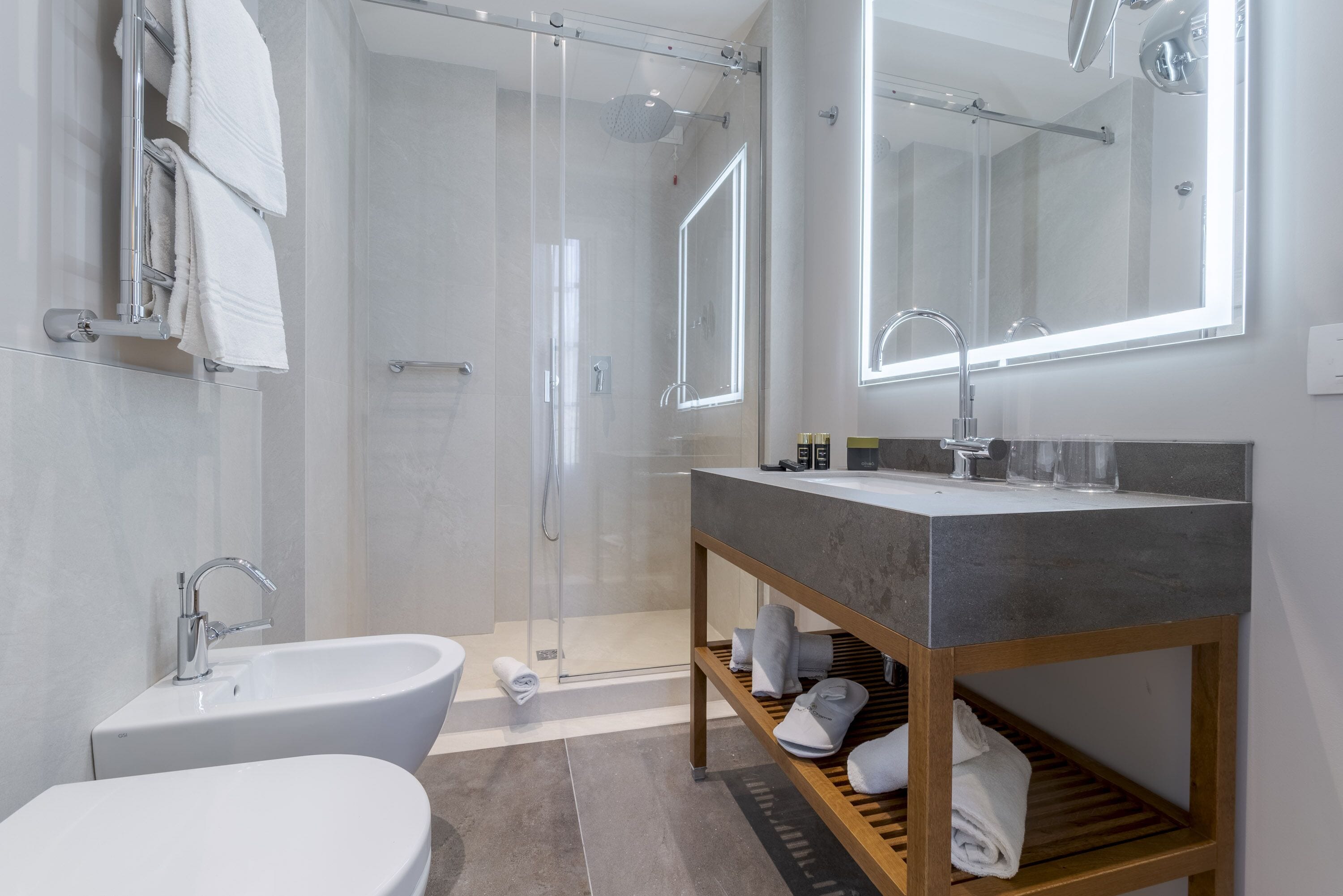 Un baño moderno con bañera, cabina de ducha con mampara de vidrio, tocador con espejo y estante de madera con toallas.