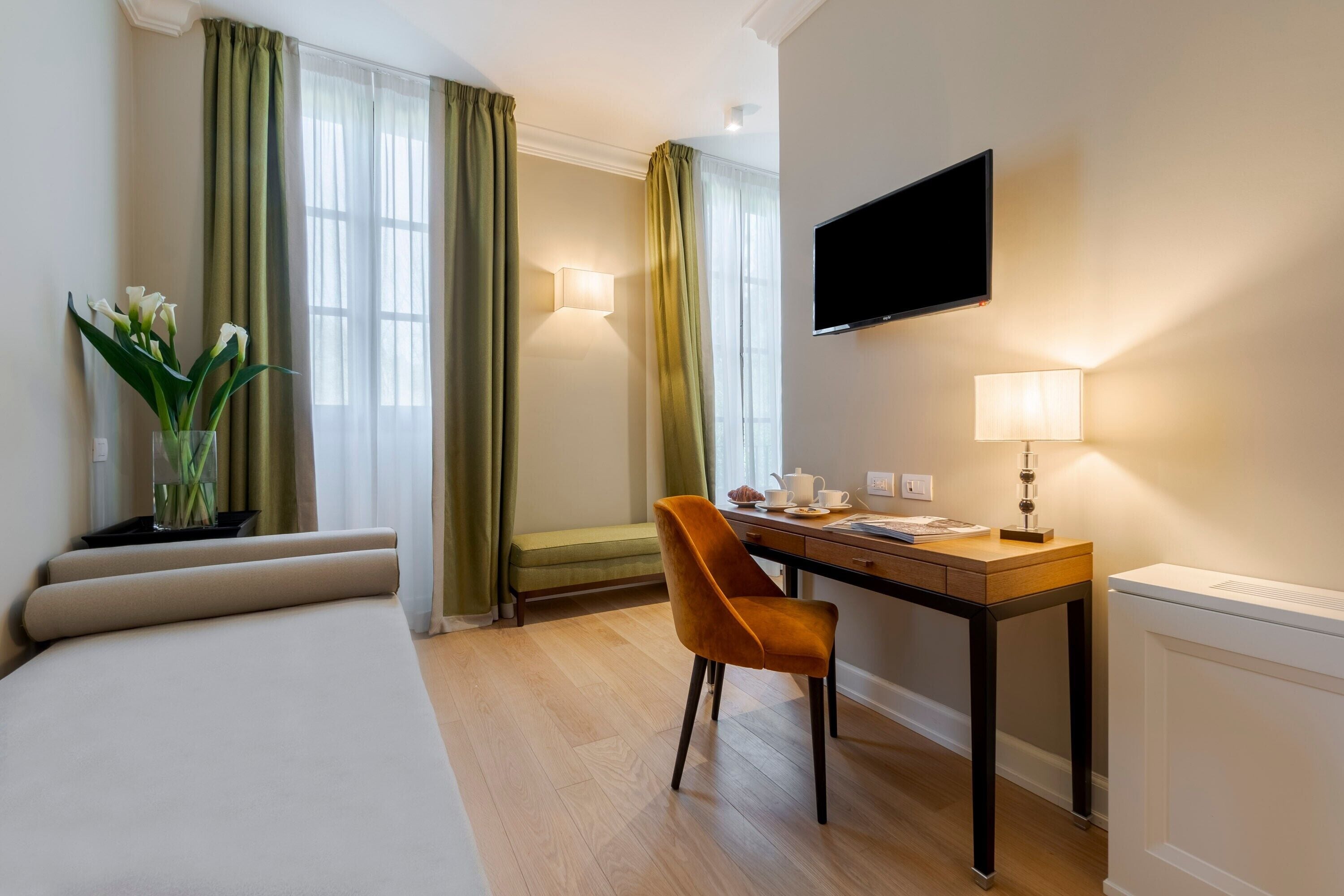 A hotel room with a bed, a desk with a chair, a TV, a lamp, and a vase with flowers.