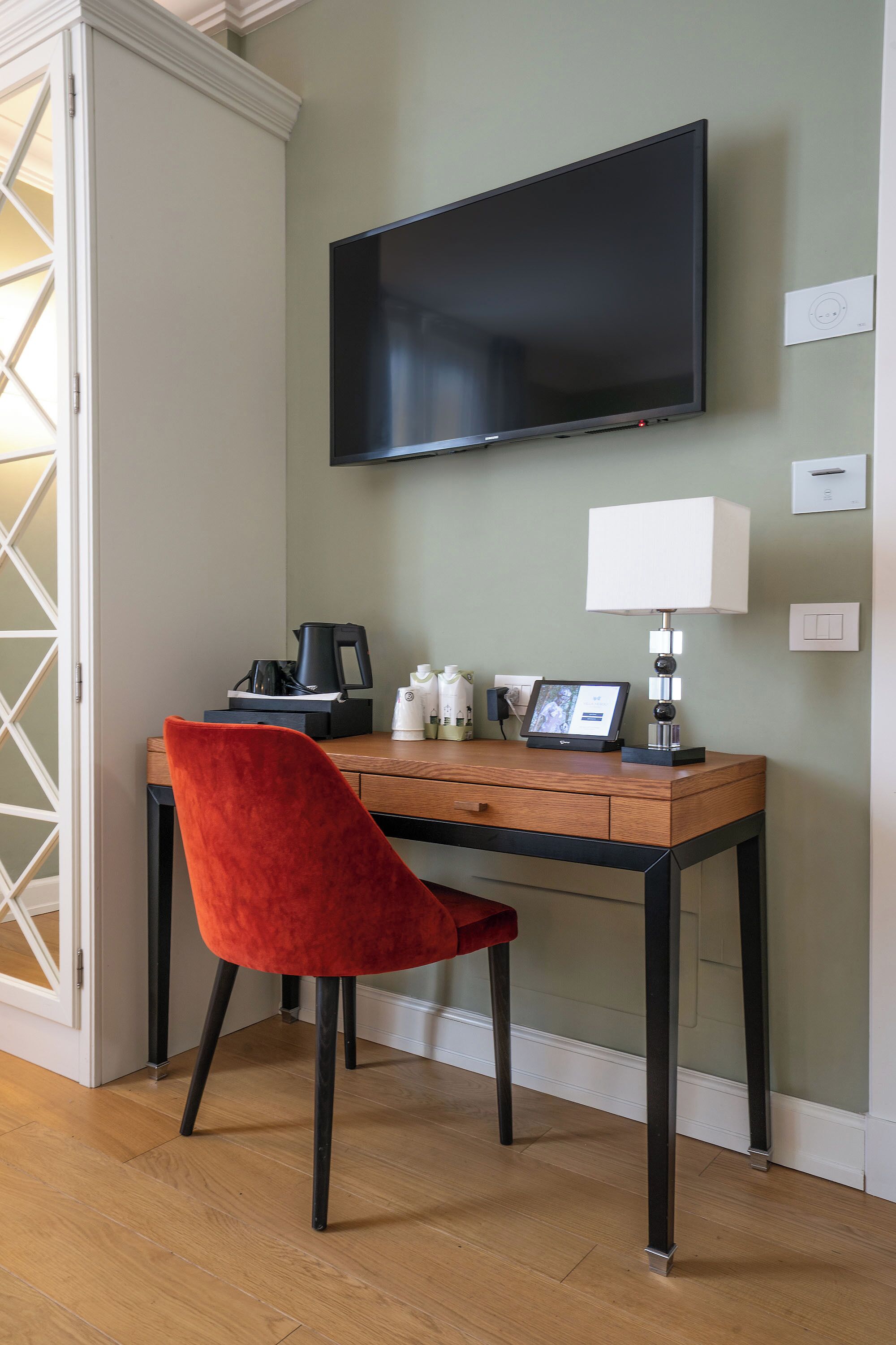 A room with a desk, a red chair, a flat-screen TV, a lamp, and a coffee maker.
