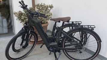 A stationary electric bicycle with a brown seat and a rear rack, parked on a tiled surface next to a white wall and a potted plant.