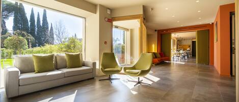 Una sala de estar moderna con un sofá y dos sillones, ventanas amplias y vista a árboles.