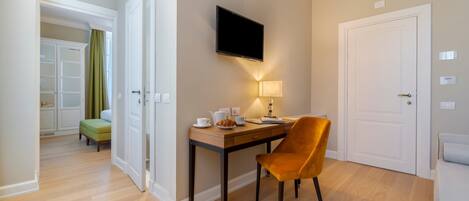 A hotel room with a wooden floor, a desk with a chair, a flat-screen TV mounted on the wall, a bedside table with a lamp, and a door.