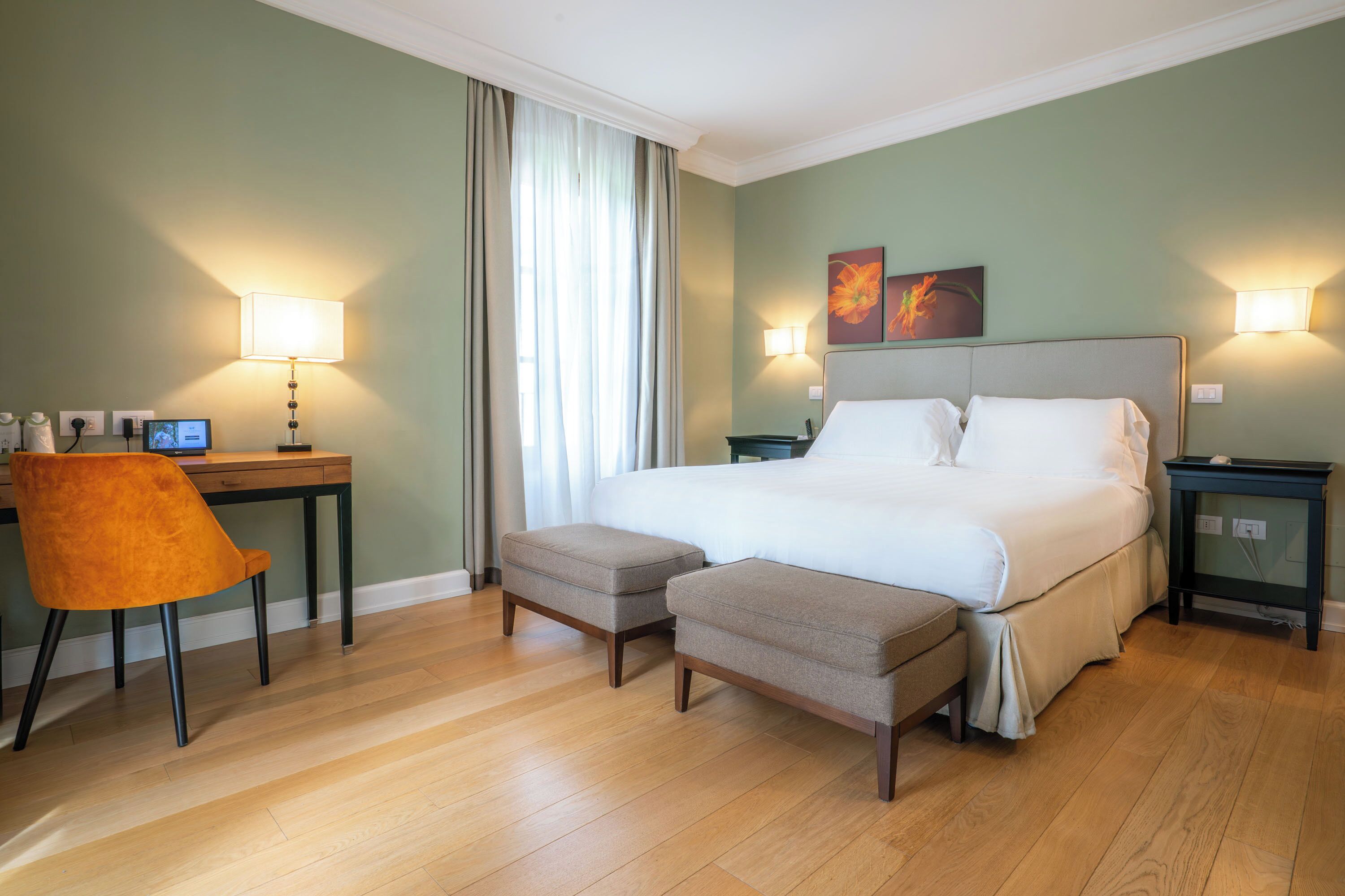 A hotel room with a bed, two bedside tables, a desk with a chair, and a window with curtains.