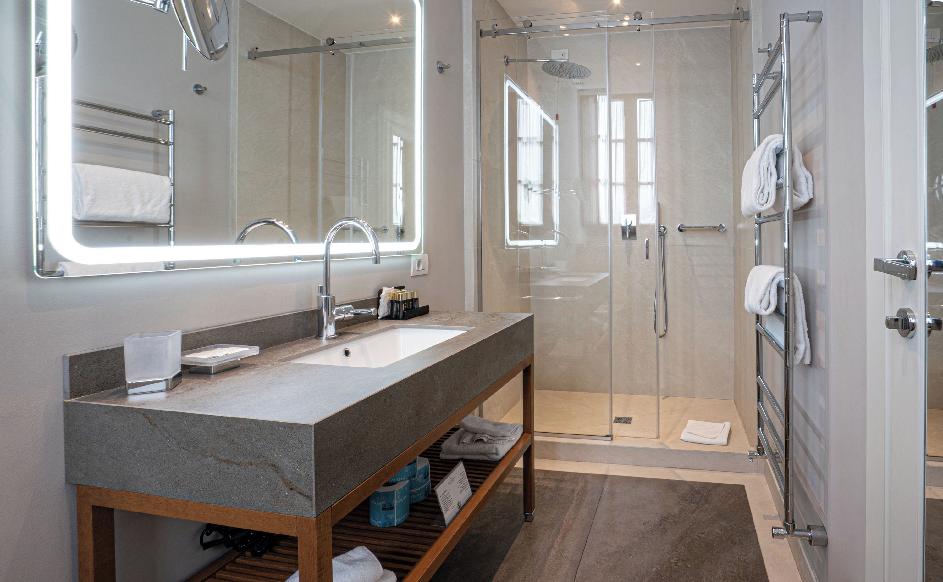 Et moderne badeværelse med vask af granit, et stort spejl, bruseafskærmning i glas og en trækommode.