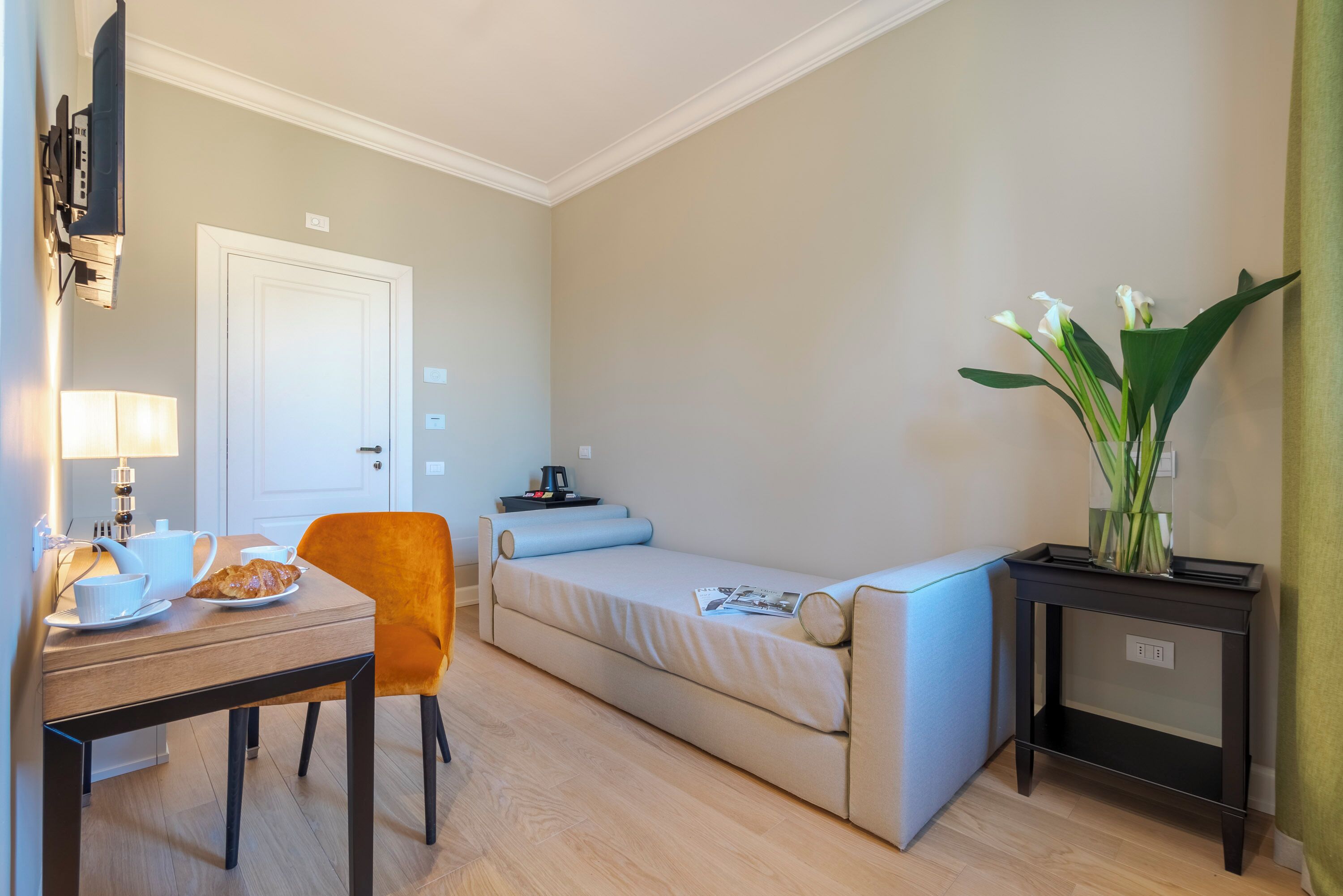 A compact room with a bed, a small table with a chair, a lamp, and a vase with flowers.