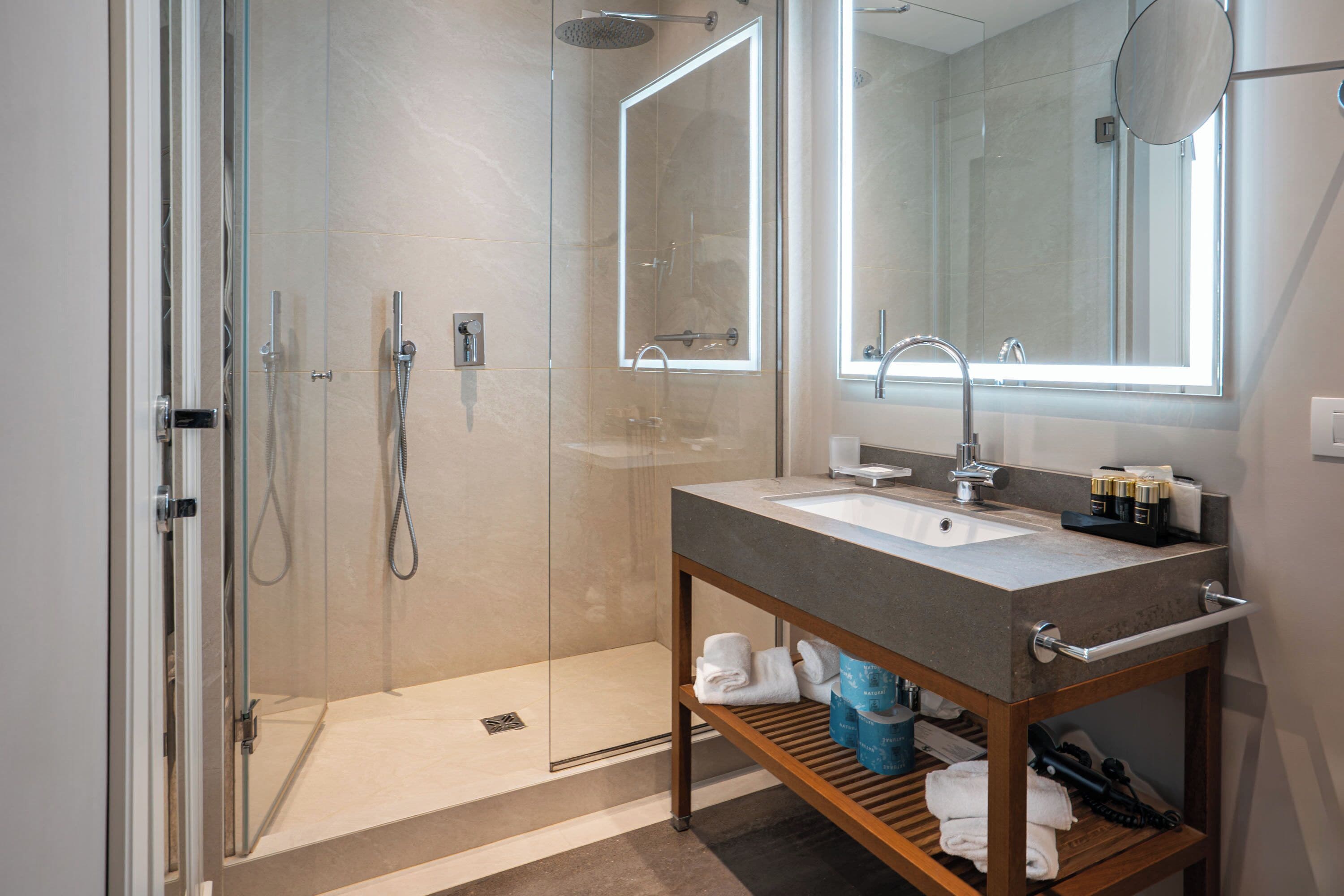 Ein modernes Badezimmer mit einer Glaskabine zur Dusche, einem Waschbecken mit Spiegel darüber und einer Holzablage mit Handtüchern und Toilettenartikeln.