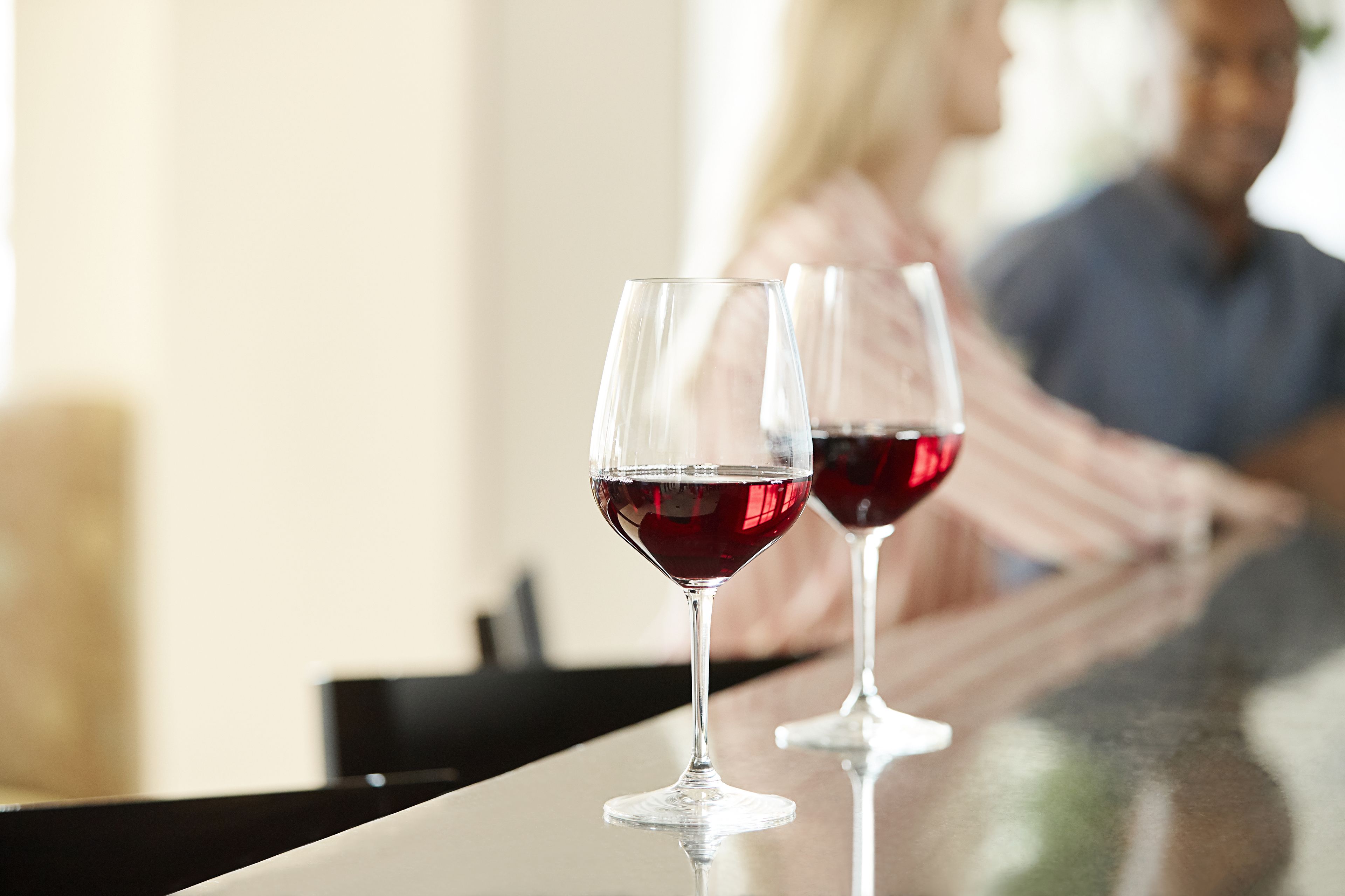 Two wine glasses on a reflective surface, likely a bar or counter, with a blurred background suggesting a social setting.